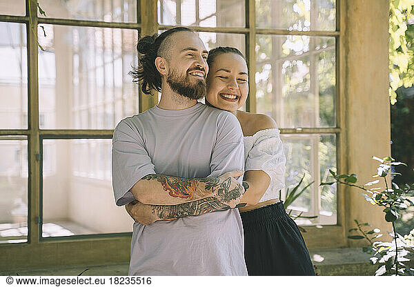 Happy woman embracing hipster boyfriend from behind in front of window