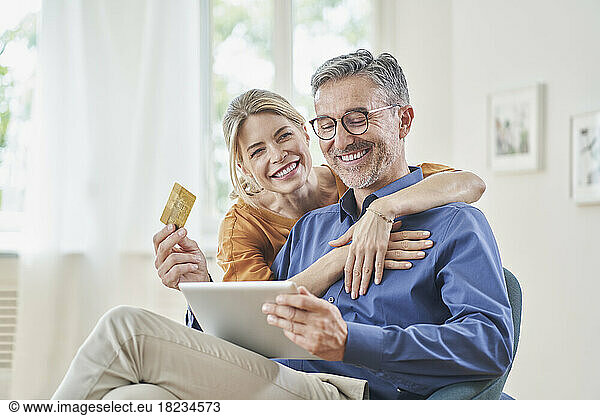 Happy woman embracing man with credit card doing online shopping through tablet PC at home
