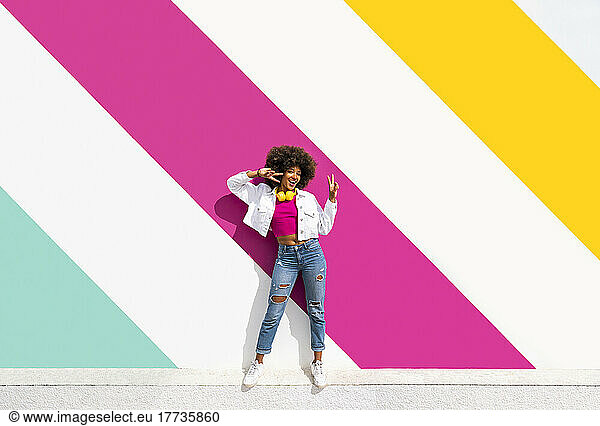Happy woman gesturing peace sign in front of colorful wall