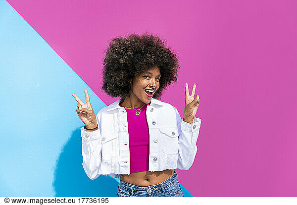 Happy woman gesturing peace sign in front of pink and blue wall