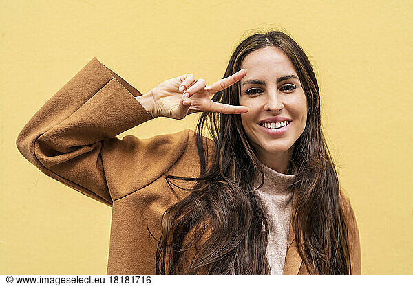 Happy woman gesturing peace sign in front of yellow wall