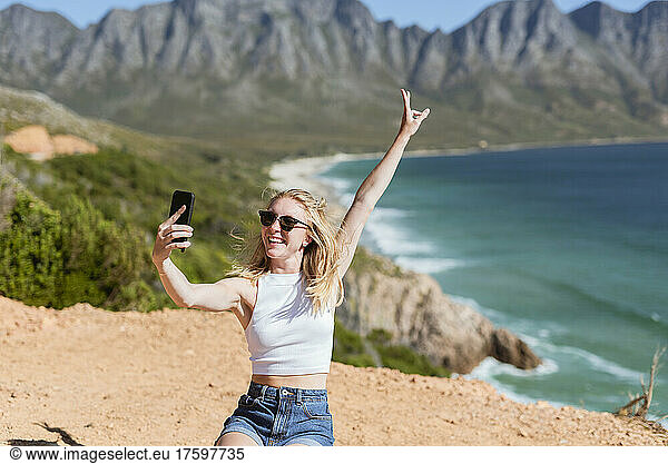 Happy woman gesturing peace sign taking selfie through mobile phone on sunny day