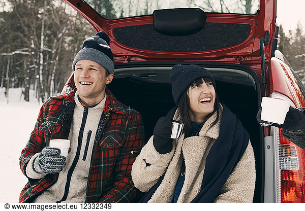 Happy woman having coffee while sitting with friends in car trunk during winter