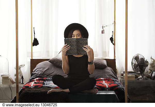Happy woman hiding face with book while sitting on bed at home