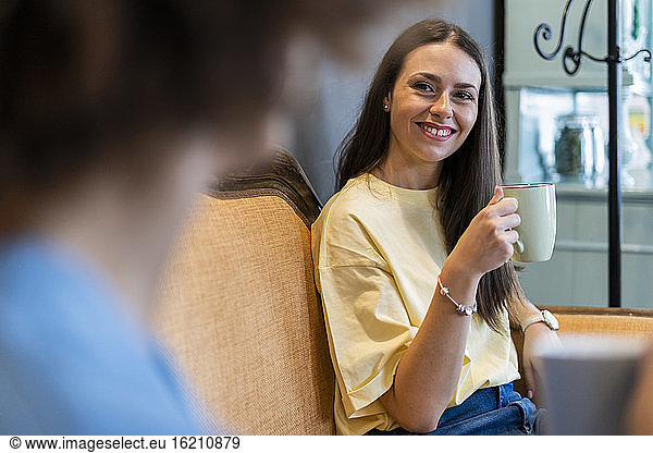 Happy woman holding coffee mug while looking friend in coffee shop