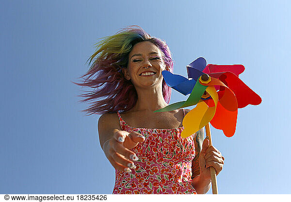 Happy woman holding colorful pinwheel toy gesturing under blue sky