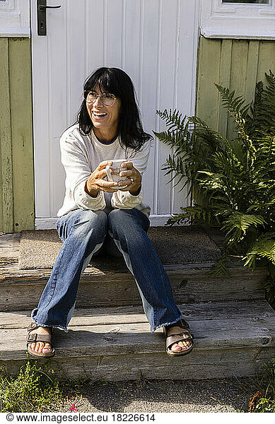 Happy woman holding cup looking away while sitting on porch outside house