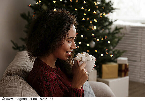 Happy woman holding cup of marshmallow cocoa sitting on sofa at home
