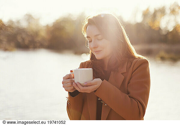 Happy woman holding cup of tea near lake
