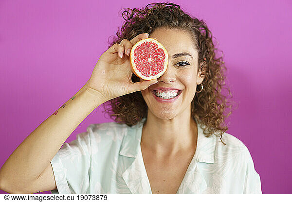 Happy woman holding grapefruit over eye against magenta background
