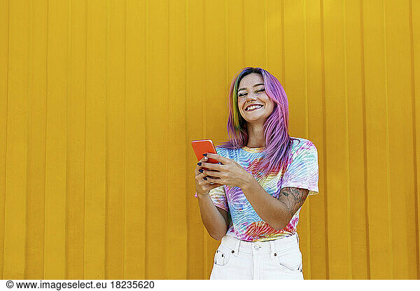 Happy woman holding mobile phone in front of yellow wall