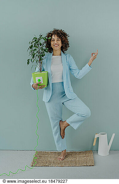 Happy woman holding potted plant connected with electric plug in front of wall