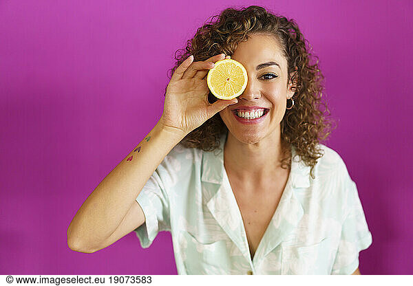 Happy woman holding slice of lemon over eye against magenta background
