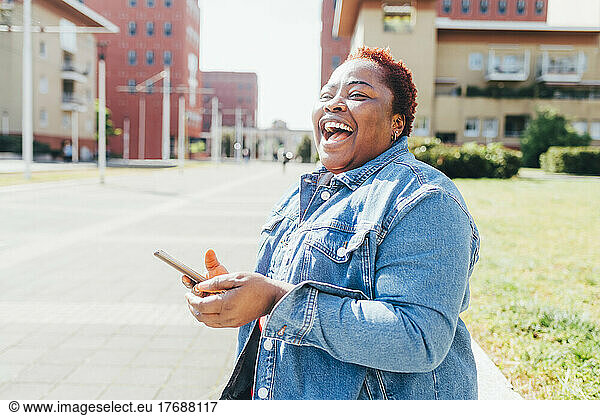 Happy woman holding smart phone laughing in the street