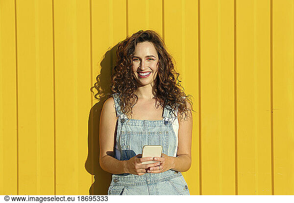 Happy woman holding smart phone standing in front of yellow wall