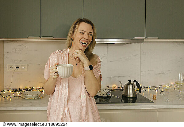 Happy woman holding tea cup standing in kitchen at home