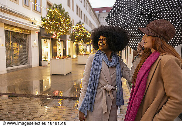 Happy woman holding umbrella and standing with friend at footpath