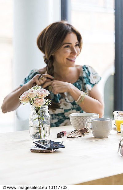Happy woman looking away while braiding hair at cafe with flower vase and smart phones on table