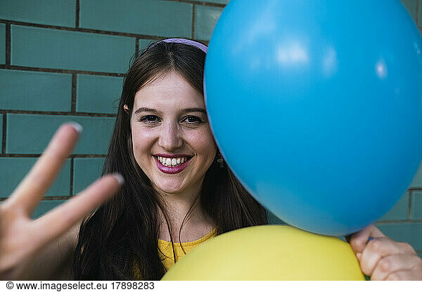 Happy woman making peace sign holding blue and yellow balloons