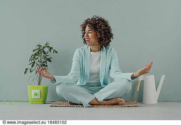 Happy woman meditating by potted plant and watering can in front of blue wall