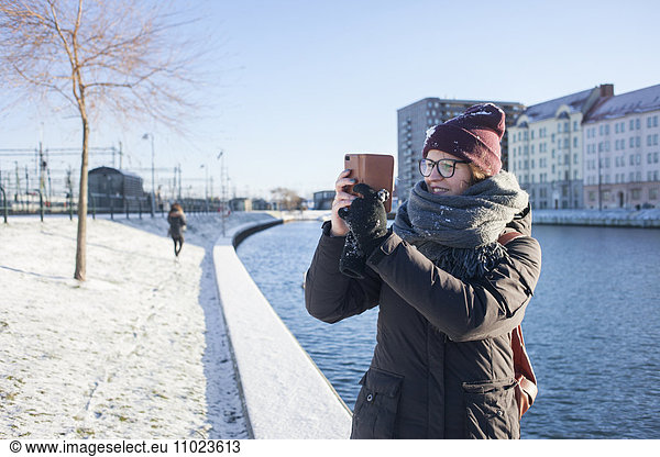Happy woman photographing through smart phone by canal in city during winter