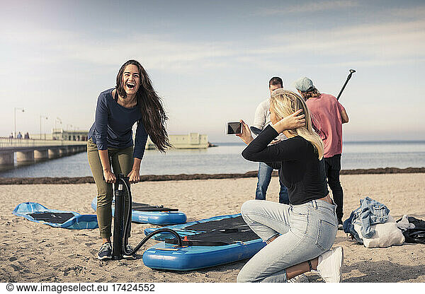 Happy woman pumping paddleboard while female friend photographing at beach