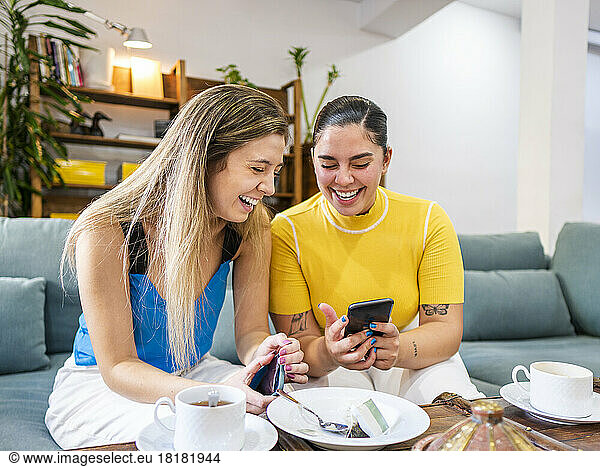 Happy woman sharing mobile phone with friend on sofa at home