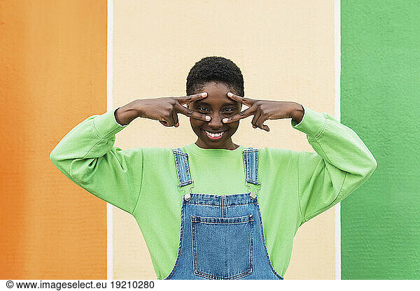 Happy woman showing peace sign gesture and touching face in front of multi colored wall