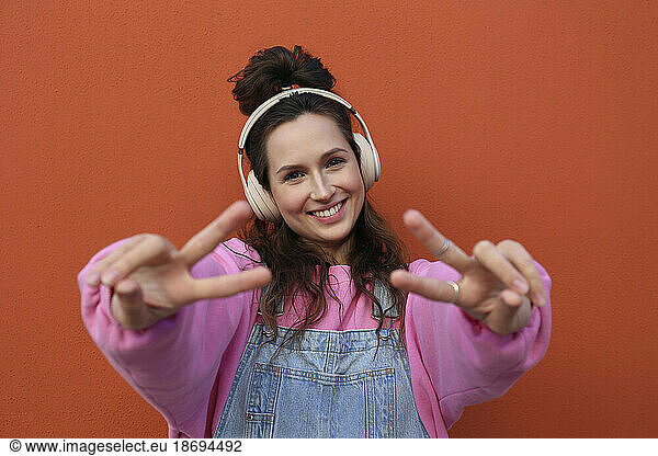 Happy woman showing peace sign wearing headphones against orange background