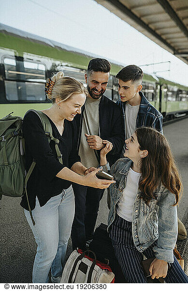 Happy woman showing smart phone to family while waiting for train at railroad station