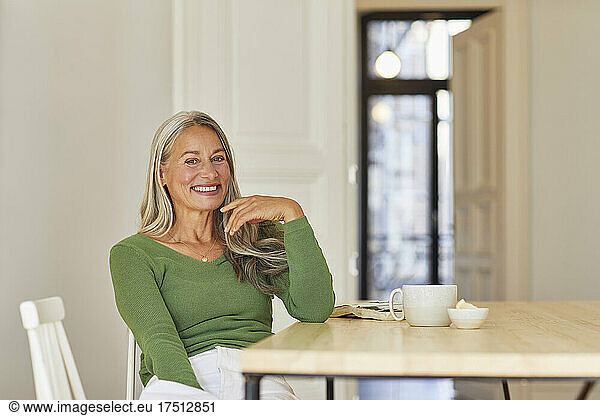 Happy woman sitting by book and cup over table at home