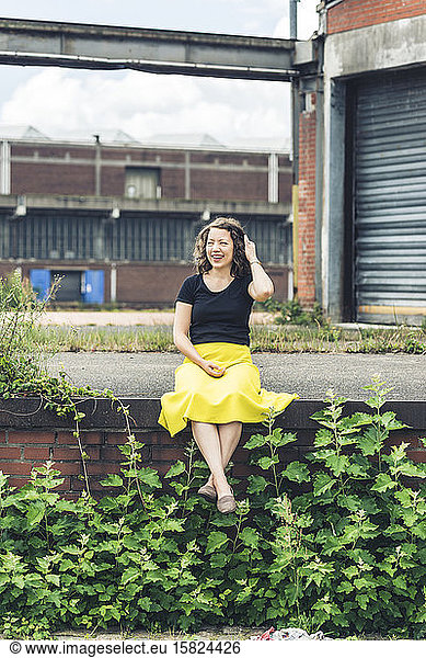 Happy woman sitting on a plant covered wall in an old industrial area