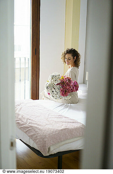 Happy woman sitting with bunch of flowers on bed