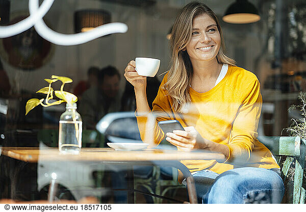 Happy woman sitting with coffee cup in cafe seen through glass