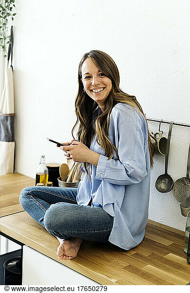Happy woman sitting with cross-legged on kitchen counter at home