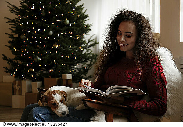 Happy woman sitting with dog and reading book at home