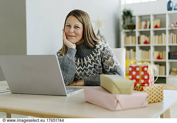 Happy woman sitting with laptop and Christmas presents on table at home