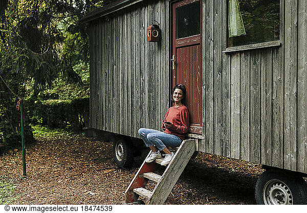 Happy woman sitting with smart phone on steps in front of cabin