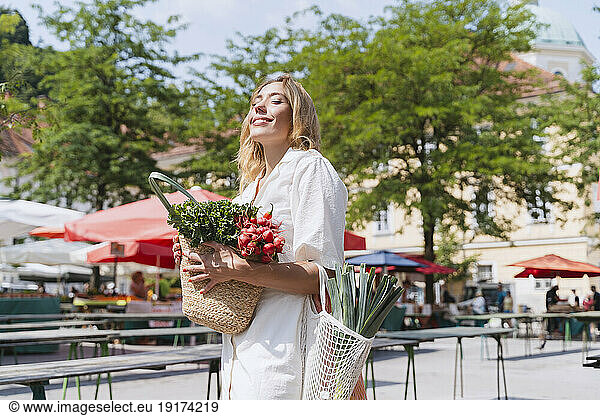 Happy woman standing with bags of groceries in farmer's market on sunny day