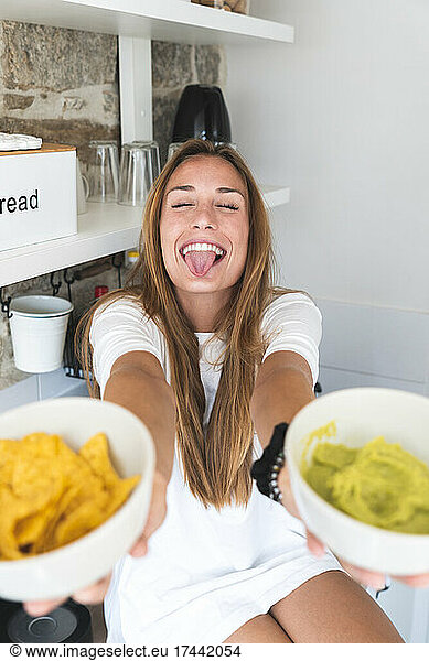 Happy woman sticking out tongue while offering nacho chips and guacamole in kitchen