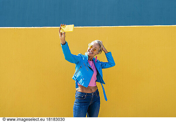 Happy woman taking selfie through mobile phone in front of colored wall