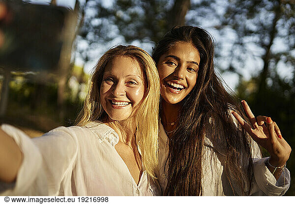 Happy woman taking selfie with teenage daughter showing peace sign in forest