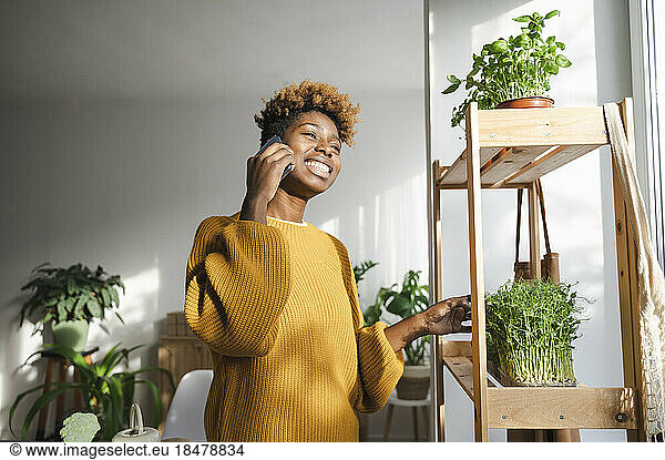 Happy woman talking on smart phone standing by plant shelf