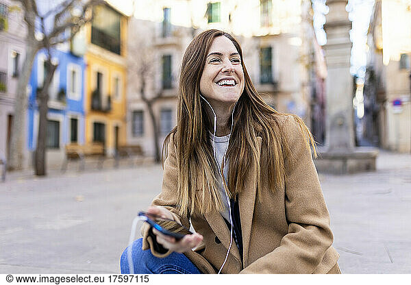Happy woman talking on smart phone through in-ear headphones at city street