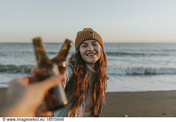 Happy woman toasting beer bottle with friend in front of sea