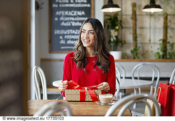 Happy woman tying ribbon of Christmas present sitting at table in cafe