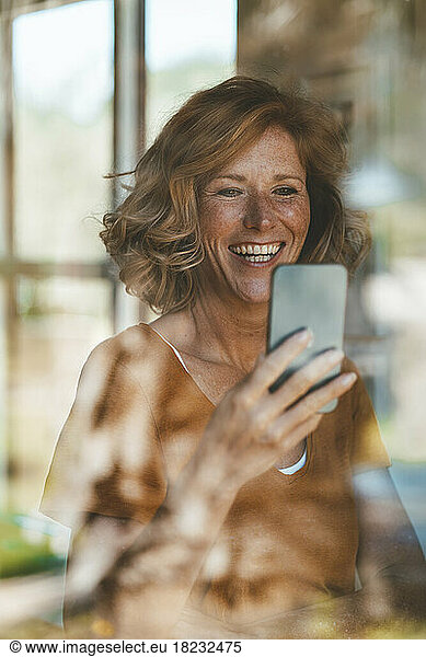 Happy woman using mobile phone seen through glass window