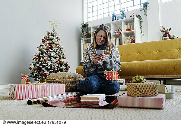 Happy woman using mobile phone sitting by gifts at home
