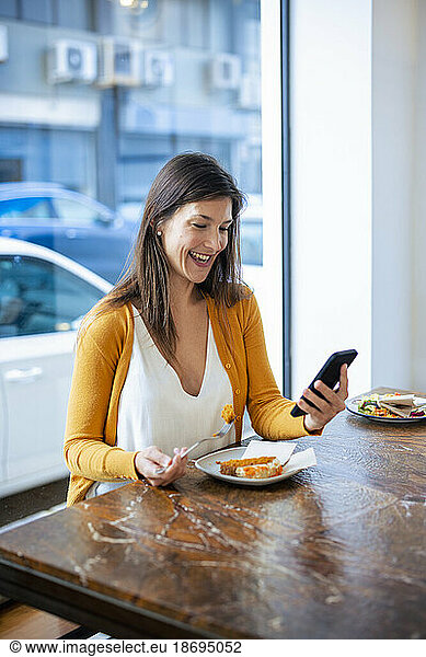 Happy woman using smart phone sitting with food on table at cafe