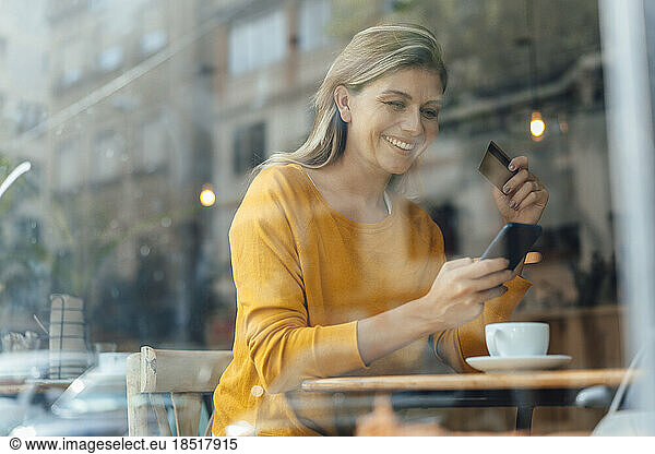 Happy woman using smart phone with credit card in cafe seen through glass